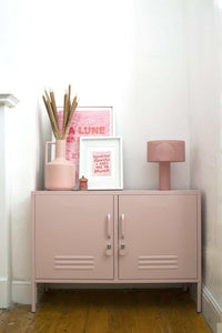 Blush pink Mustard Made locker console with two doors, metal handles, placed in a cozy room corner with a pink vase, framed art, and a pink lamp on top