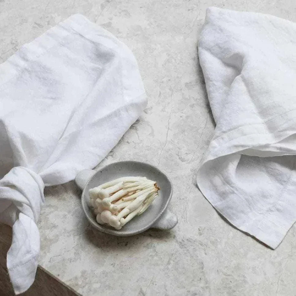 Set of four white Rendr 100% linen napkins displayed on a marble surface with a small dish of mushrooms.