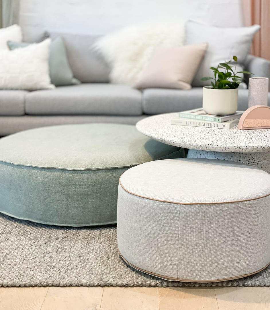Soft round custom ottomans upholstered in neutral Warwick fabric with blush leather piping, placed in a modern living room setting.