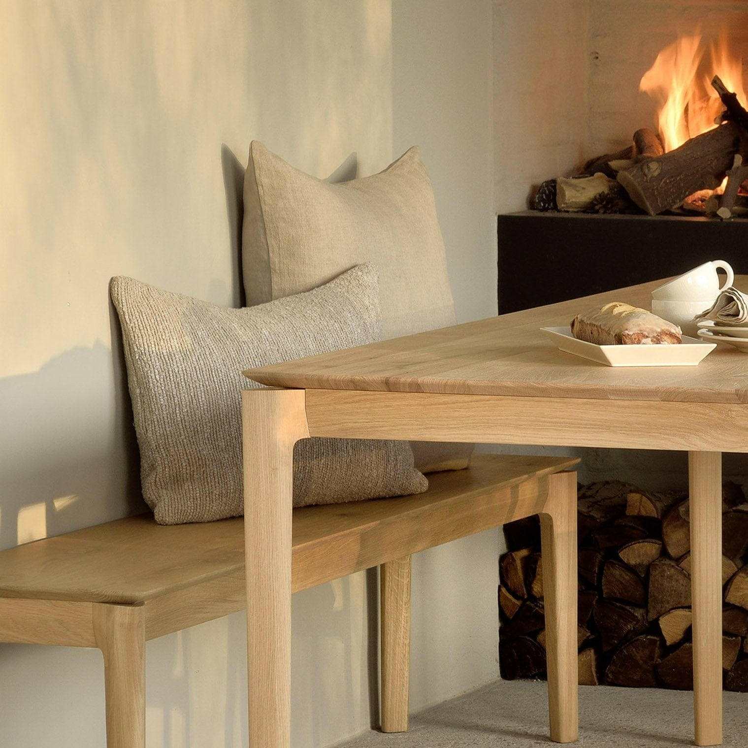 Ethnicraft Bok Dining Table in natural oak with smooth edges and sturdy legs, paired with a wooden bench and cushions near a cozy fireplace.