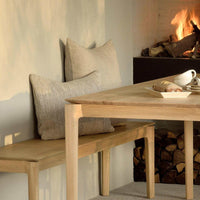 Ethnicraft Bok Dining Table in natural oak with smooth edges and sturdy legs, paired with a wooden bench and cushions near a cozy fireplace.