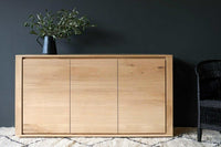 Ethnicraft Oak Shadow Sideboard made of natural solid European oak with three doors, placed against a dark wall with a plant on top and a black wicker chair beside it.