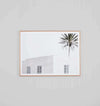 Minimalist photographic print of a white desert villa with a palm tree, framed in oak.