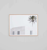 Minimalist photographic print of a white desert villa with a palm tree, framed in oak.