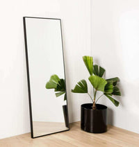 Middle of Nowhere Flynn Leaner Mirror in black with minimalist wooden frame leaning against white wall next to potted green plant
