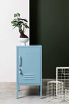 Blue Mustard Made Locker The Shorty Ocean with handle, lock, and ventilation vents standing next to a green wall and wire baskets.