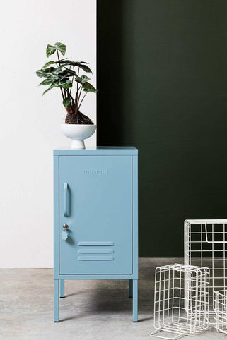 Blue Mustard Made Locker The Shorty Ocean with handle, lock, and ventilation vents standing next to a green wall and wire baskets.
