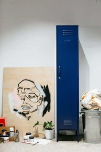 Slim navy blue Mustard Made Locker standing against a wall beside art supplies and a painted canvas.