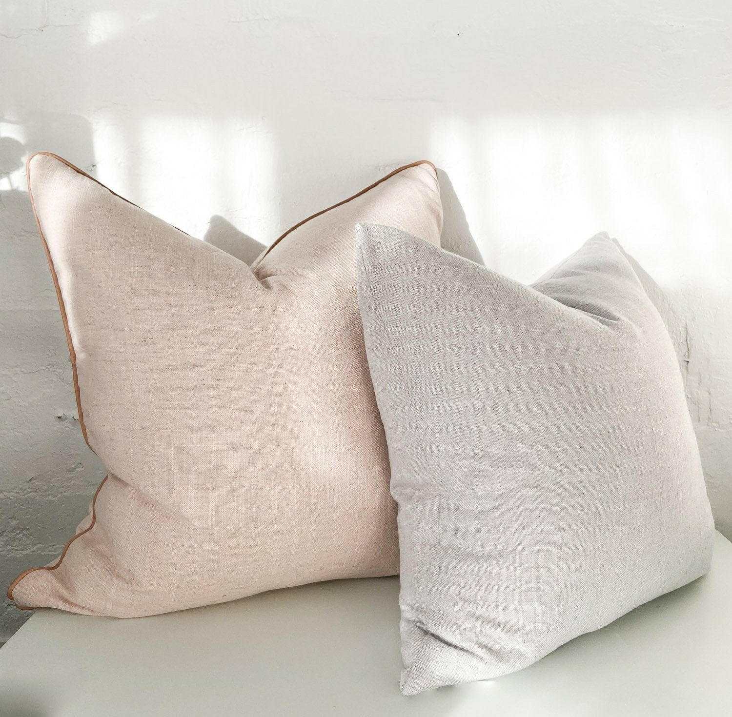 Two norsu Studio Cushions in soft beige and light gray, featuring blush leather piping on the beige cushion, displayed on a white surface against a white wall.