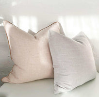 Two norsu Studio Cushions in soft beige and light gray, featuring blush leather piping on the beige cushion, displayed on a white surface against a white wall.