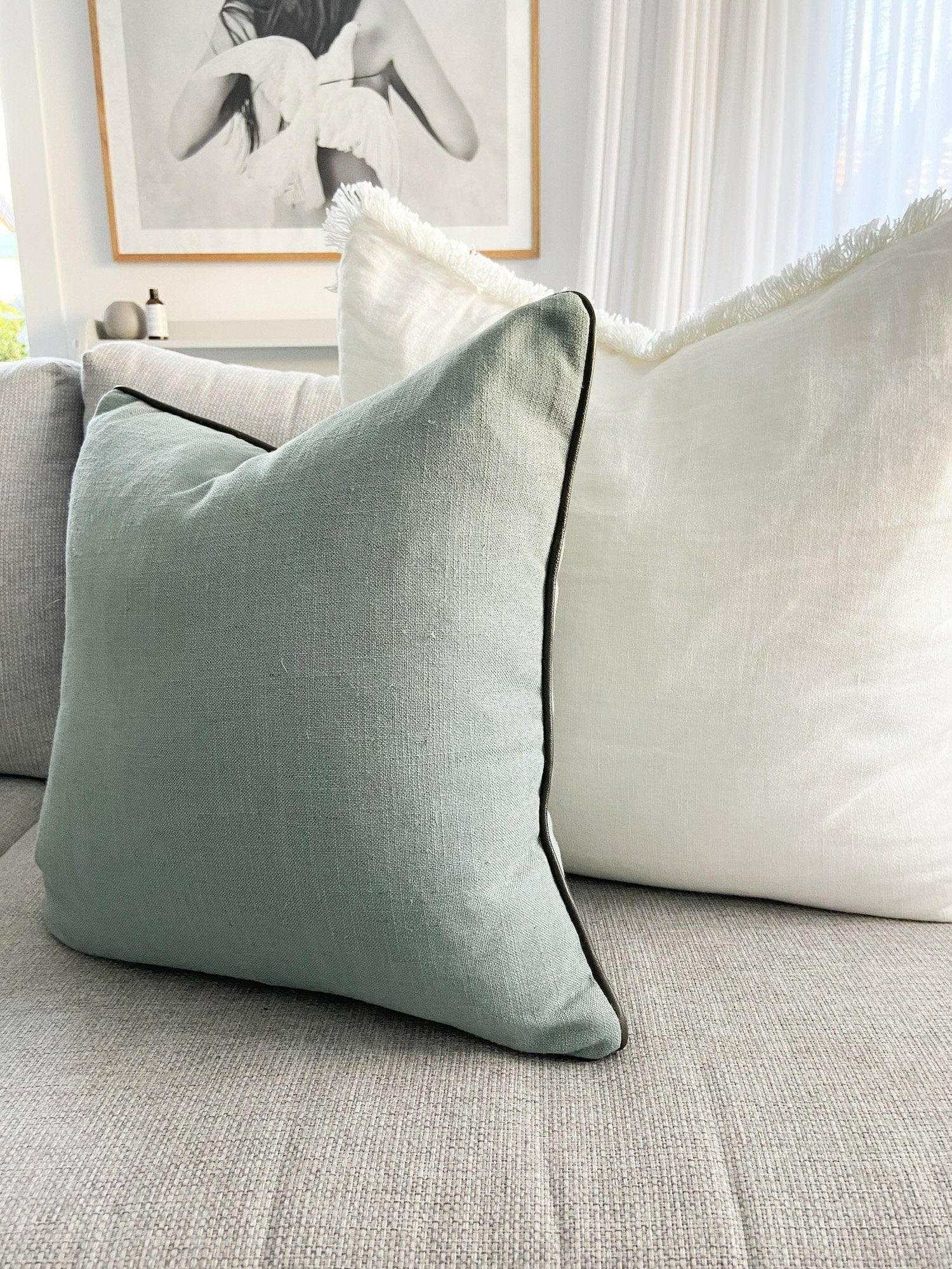 norsu cushion in Lexus Seaglass fabric with olive leather piping on a sofa with white cushions in the background