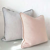 Two norsu cushions, one beige with charcoal piping and one light pink with matching piping, displayed against a white textured wall.