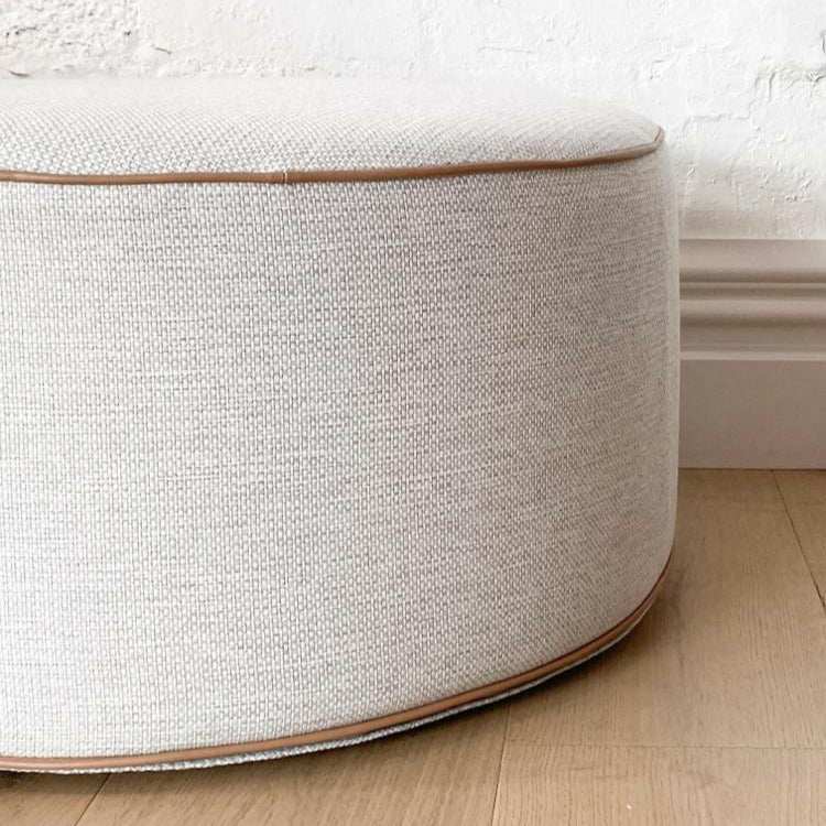 Round norsu Ottoman in Lindeman Snow fabric with blush leather piping on wooden floor against white wall