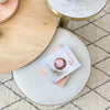 norsu Ottoman in Lindeman Snow fabric with blush leather piping, round soft ottoman with books on top on patterned carpet