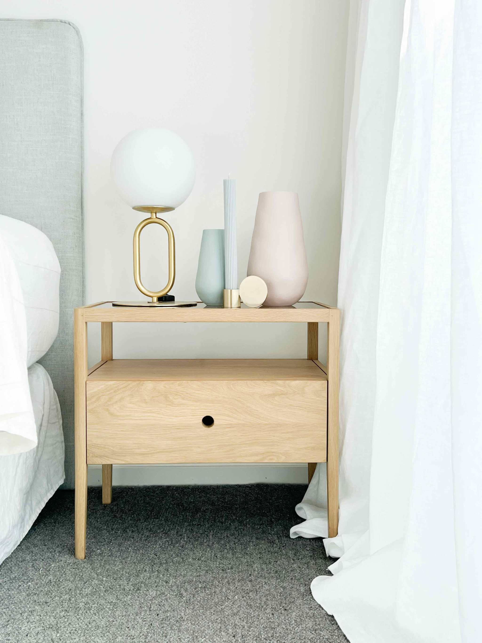 Ethnicraft Spindle Bedside table in natural oak with a single drawer and smokey glass top placed beside a bed with decorative vases and lamp.