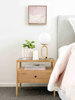 Ethnicraft Spindle Bedside table in natural oak with single drawer and smokey glass top next to bed with lamp and plant on top