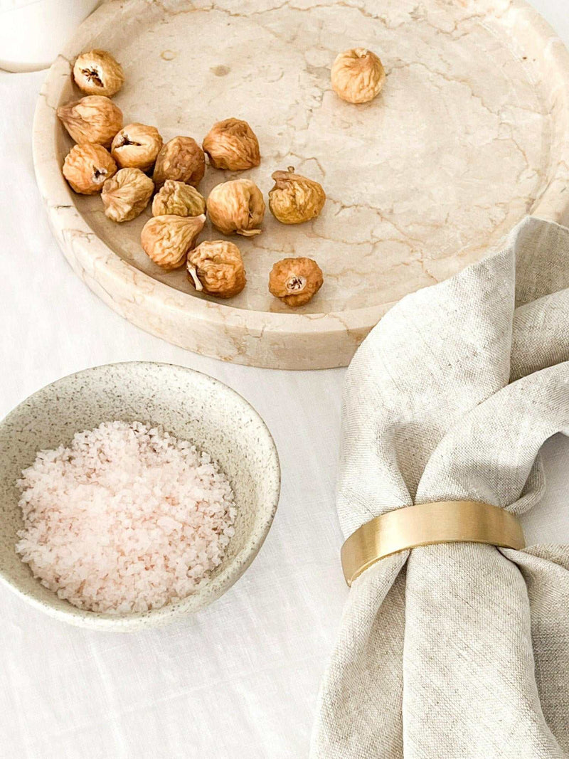 Set of four Rendr 100% linen natural napkins with a gold napkin ring beside dried figs and a bowl of pink salt on a marble tray