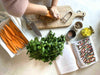 Hands chopping sweet potatoes on wooden board next to fresh herbs and open recipe book from The Beauty Chef by Carla Oates