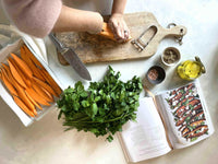 Hands chopping sweet potatoes on wooden board next to fresh herbs and open recipe book from The Beauty Chef by Carla Oates
